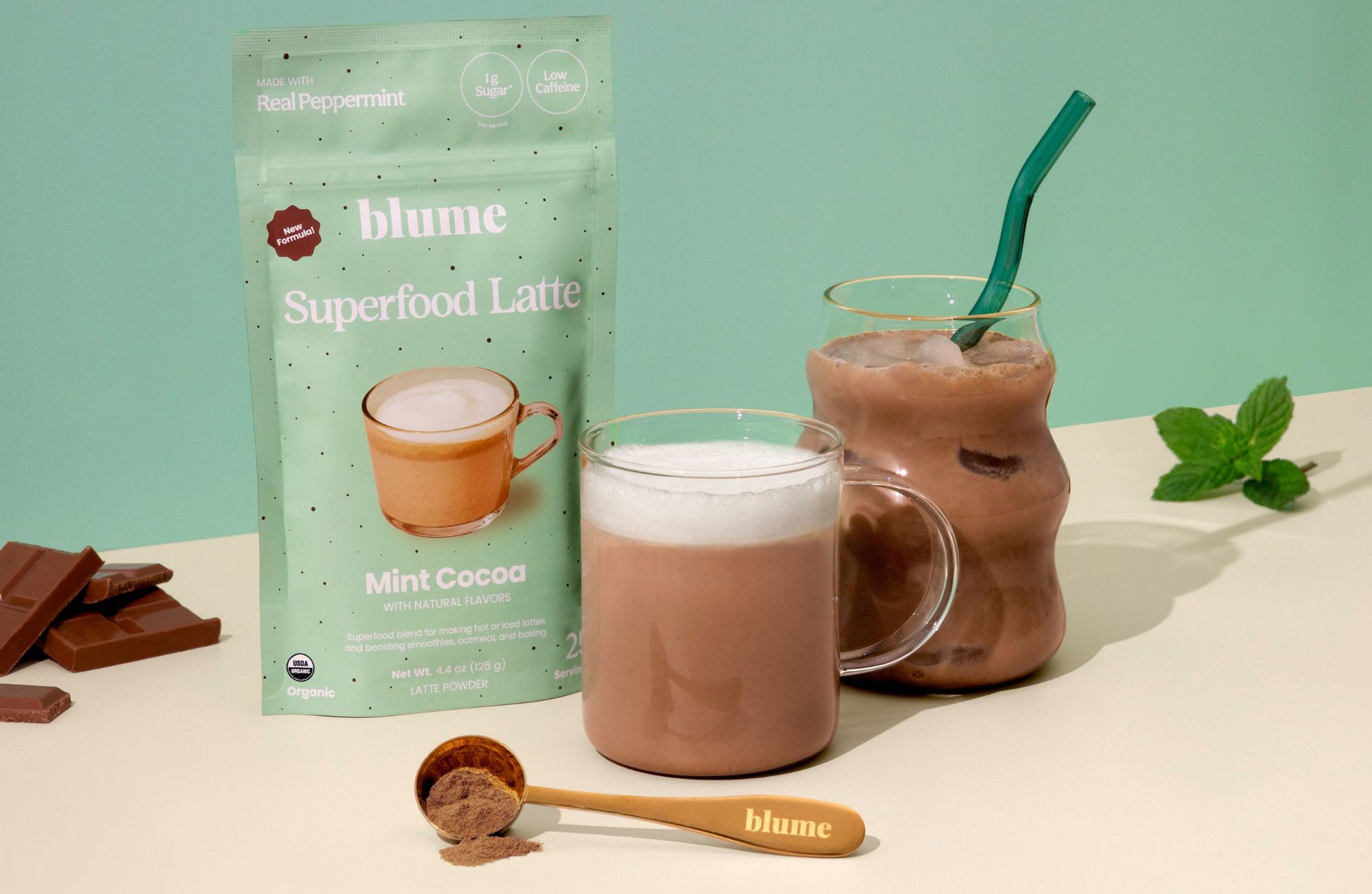 A package of Blume Mint Cocoa Superfood Latte is displayed with a glass mug of frothy cocoa, a milkshake with ice cubes and a straw, a wooden spoon of powder, chocolate pieces, and mint leaves on a light green background.